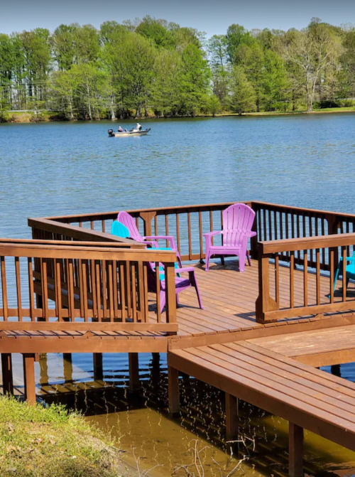 A wooden dock with colorful chairs overlooks a calm lake, with a boat in the distance and trees lining the shore.