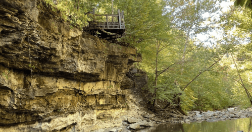 A rocky cliff with a wooden overlook surrounded by lush green trees beside a calm river.