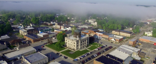 Aerial view of a small town with a courthouse, surrounded by fog and greenery, showcasing buildings and streets.
