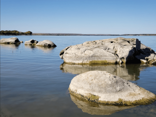 Calm water with large, smooth rocks emerging from the surface under a clear blue sky.