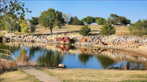 A serene park scene featuring a calm pond, rocky shoreline, and lush green trees under a clear blue sky.