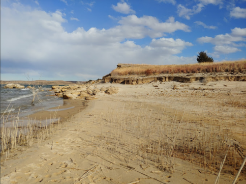 A sandy beach with sparse grass, rocky cliffs, and a cloudy blue sky in the background.