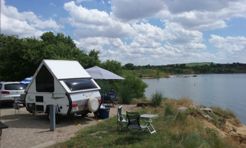 A small camper parked by a lake, with chairs set up nearby and a cloudy sky overhead.