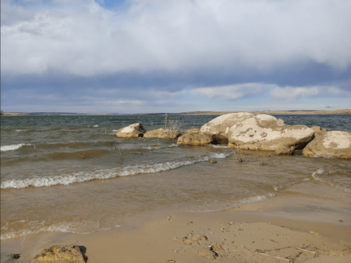 A serene lakeshore with gentle waves, sandy beach, and large rocks under a cloudy sky.