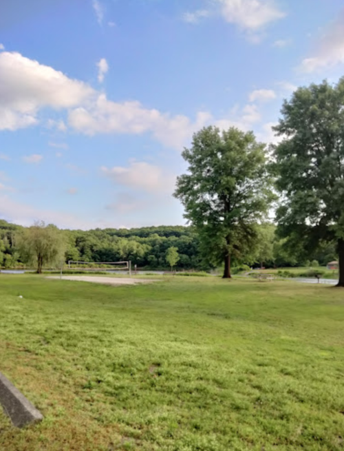 A serene park scene featuring green grass, trees, and a calm lake under a partly cloudy sky.