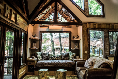 Cozy cabin interior with large windows, wooden accents, and a comfortable seating area surrounded by trees.