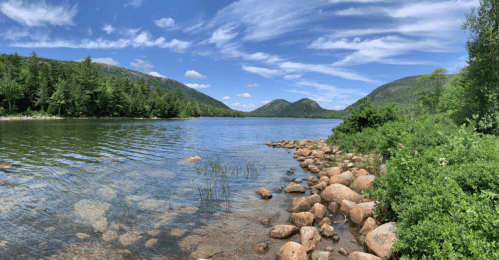 A serene lake surrounded by green hills and rocky shores under a blue sky with fluffy clouds.