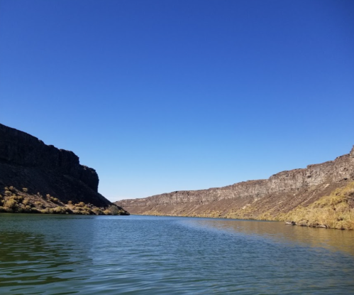 A serene river flows between steep, rocky cliffs under a clear blue sky.