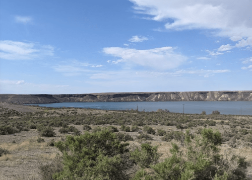 A serene landscape featuring a calm lake surrounded by rocky cliffs and sparse vegetation under a blue sky.