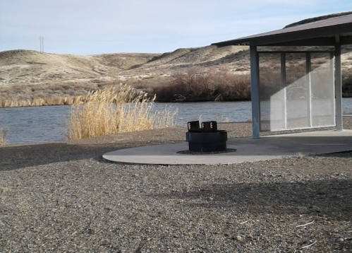A riverside scene featuring a shelter, a fire pit, and dry grasslands in the background under a clear sky.