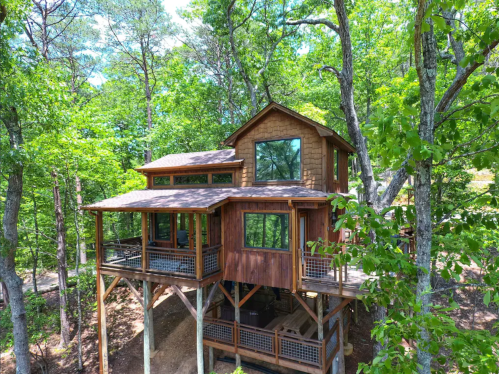 A wooden treehouse nestled among lush green trees, featuring multiple levels and large windows.