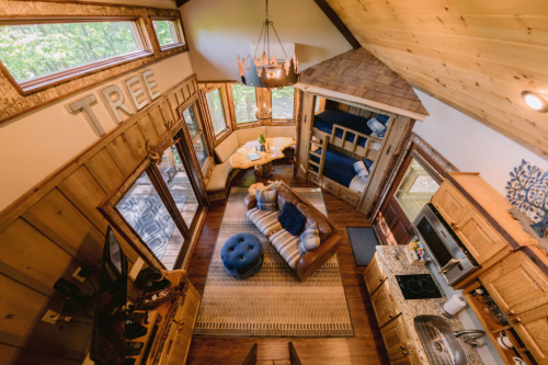 Cozy wooden interior of a treehouse with a living area, kitchen, and lofted sleeping space, surrounded by large windows.