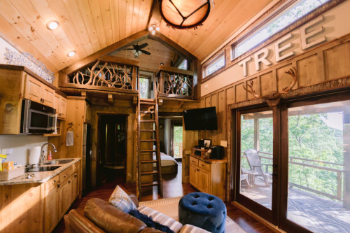 Cozy wooden cabin interior with a loft, kitchen, and large windows overlooking nature. Rustic decor and warm lighting.