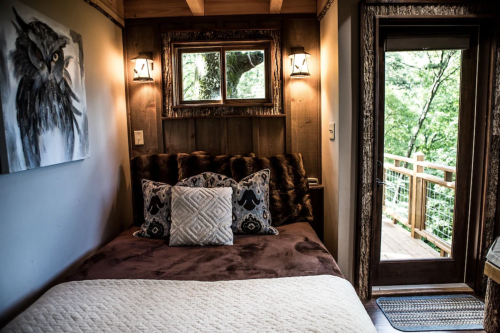 Cozy bedroom with a plush bed, decorative pillows, and a door leading to a forest view. Warm wooden accents throughout.