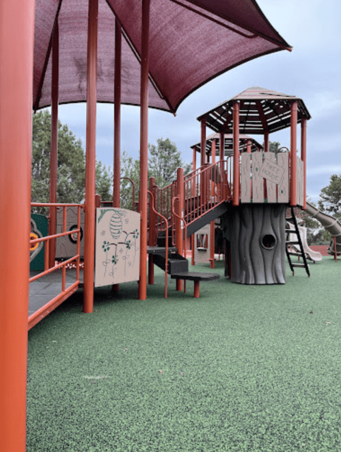 Colorful playground structure with slides and climbing areas, surrounded by green rubber flooring and shaded by canopies.