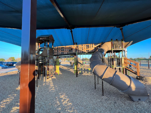 Playground structure with a large slide, shaded by a blue canopy, surrounded by gravel and open space.