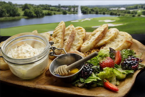 A wooden platter with bread, a jar of creamy dip, honey, and a fresh salad with berries, set against a scenic golf course view.