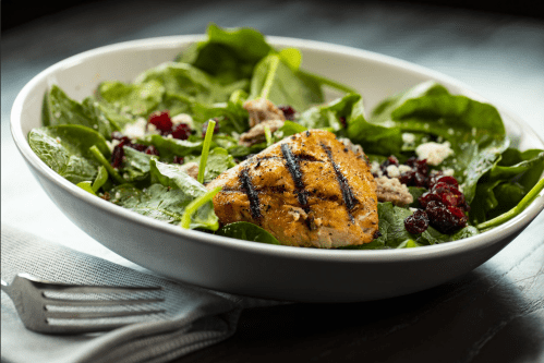 A bowl of spinach salad topped with grilled salmon, cranberries, and feta cheese, served with a fork and napkin.