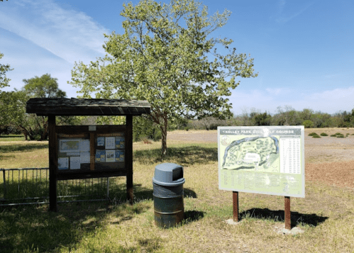 A park information board and a map sign in a grassy area with trees under a clear blue sky.