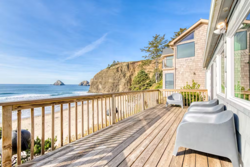 A wooden deck overlooking a beach with gentle waves and cliffs in the background under a clear blue sky.