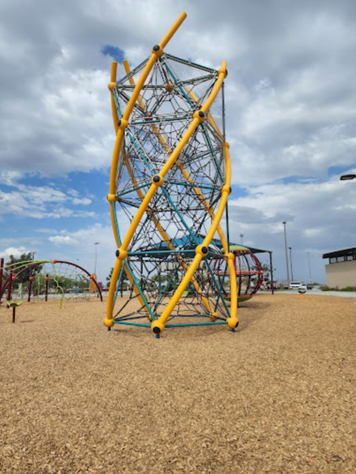 A colorful climbing structure in a playground, surrounded by wood chips and under a cloudy sky.