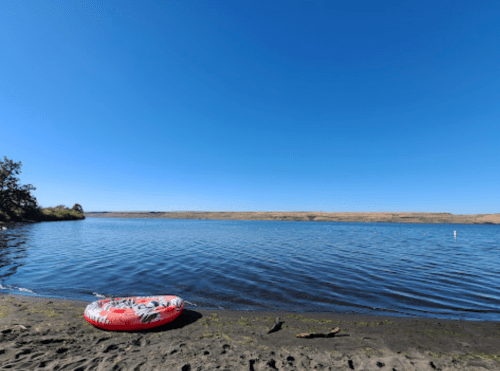 A calm river scene with a red inflatable ring on the sandy shore under a clear blue sky.