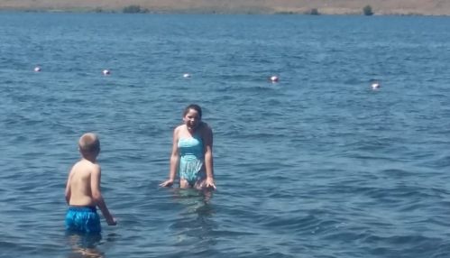 Two children play in a lake, one wearing a blue swimsuit and the other in blue shorts, enjoying a sunny day.