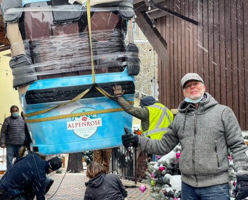 A man poses with a thumbs-up as workers lift a large hot tub in snowy conditions outside a building.