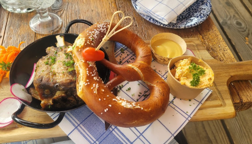 A hearty meal featuring a pretzel, meat dish, mustard, and a creamy dip, served on a wooden board with garnishes.