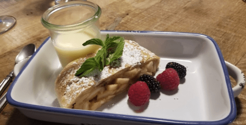 A slice of apple strudel topped with powdered sugar, mint, and served with a jar of custard and fresh berries.