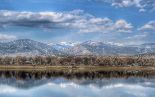 A serene lake reflects snow-capped mountains and a cloudy sky, with trees and cabins lining the shore.