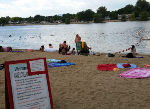 A sandy beach by a lake with people swimming and relaxing, featuring a sign with rules for visitors.