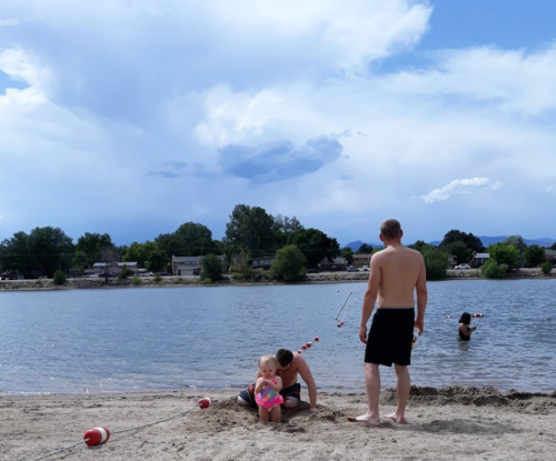 A sunny beach scene with two adults and a child playing in the sand near a lake, with clouds in the background.
