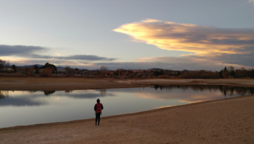 A person stands on a sandy shore, gazing at a calm lake reflecting a colorful sunset and clouds.