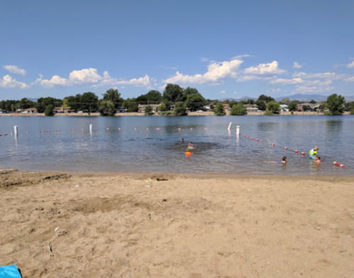 A sandy beach by a calm lake with people swimming and playing in the water under a clear blue sky.