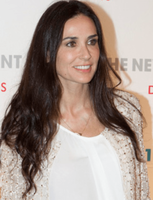 A woman with long, dark hair smiles while wearing a white top and a light, embellished jacket at an event.