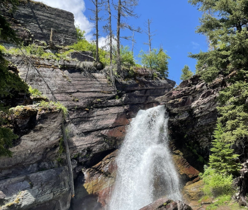 A waterfall cascades down rocky cliffs, surrounded by lush greenery and a clear blue sky.
