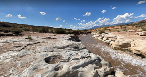 A rocky riverbank with flowing water under a blue sky dotted with clouds. Desert landscape in the background.