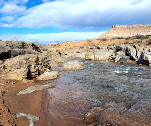 A serene river flows through rocky terrain under a partly cloudy sky, with sandy banks and distant hills in the background.