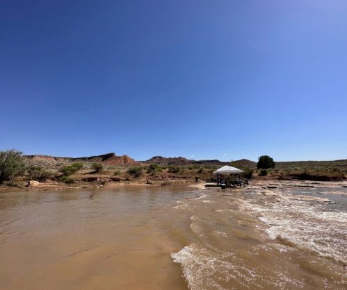 A calm river flows under a clear blue sky, with a small gazebo on the shore and rocky hills in the background.
