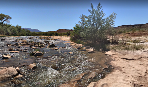 A serene river flows over rocks, surrounded by greenery and sandy banks under a clear blue sky.