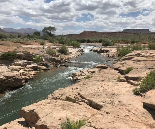 A winding river flows through rocky terrain under a cloudy sky, surrounded by sparse vegetation and distant cliffs.