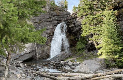 A serene waterfall cascades down rocky cliffs, surrounded by lush green trees and a tranquil stream below.