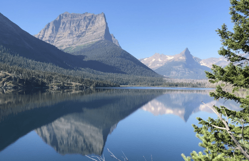 A serene lake reflects towering mountains and clear blue skies, surrounded by lush green trees.