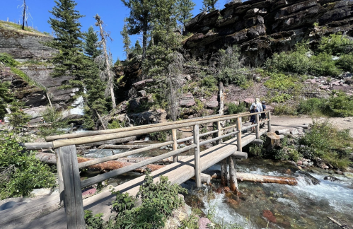 A wooden bridge crosses a clear stream surrounded by lush greenery and rocky terrain under a blue sky.