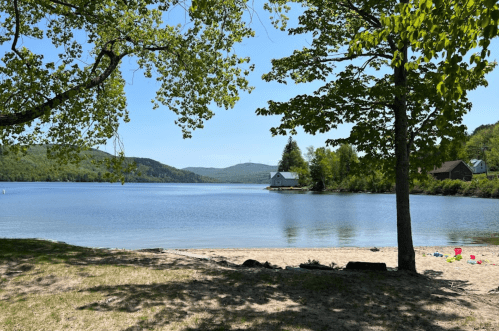 A serene lake view with sandy shore, trees, and a small house on the opposite bank under a clear blue sky.