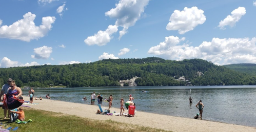A sunny beach scene with people swimming and relaxing by the water, surrounded by green hills and fluffy clouds.