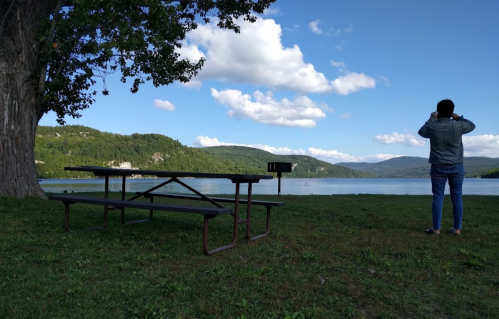 A person stands by a lake, taking photos, with a picnic table and mountains in the background under a blue sky.