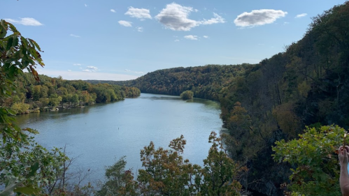 A serene river flows between lush, green hills under a clear blue sky with scattered clouds.
