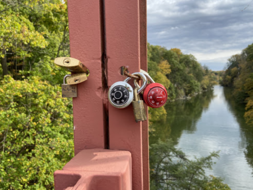 Two padlocks, one silver and one red, hang on a pink railing overlooking a calm river surrounded by trees.